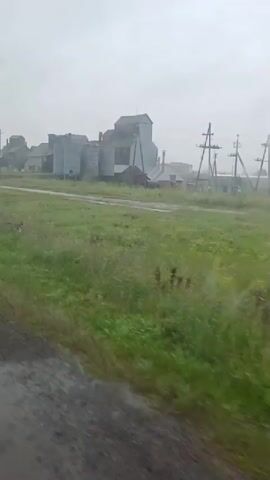 The video shows a passing view of an industrial facility with several large, gray silos and buildings. A tractor or similar vehicle is visible on the left side of the frame, and a large, cylindrical object, possibly a tank, is being transported by a vehicle in front of the buildings. A signpost with Russian text and numbers "52" and "68" is present on the left side. The environment appears to be rural with green grass and a muddy, dirt road in the foreground. The sky is overcast and the weather seems to be rainy or misty, with water droplets visible on the camera lens. The overall impression is one of a functional, perhaps somewhat aged, industrial area in a rural setting.