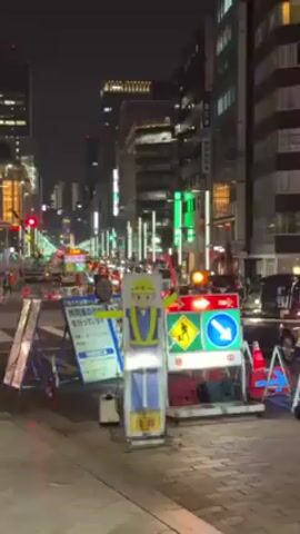 The video shows a city street at night, likely in Japan based on signage. The street has tall buildings on both sides, illuminated by streetlights and lights from the buildings. Traffic is moving through the street, with several vehicles visible. Road work is in progress, indicated by construction signs, a worker directing traffic, and traffic cones. The sky is dark.