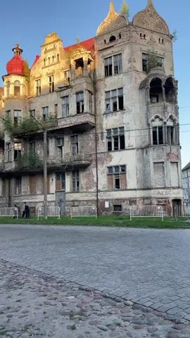 The video shows an abandoned, dilapidated building with a red roof and peeling paint. Plants are growing on the balconies and around the structure. A dark sedan drives by on a cobblestone street in front of the building. Two figures, possibly people, are visible near the front of the building, behind a white fence. The sky is clear and appears to be at sunset, casting a warm light on the building.
