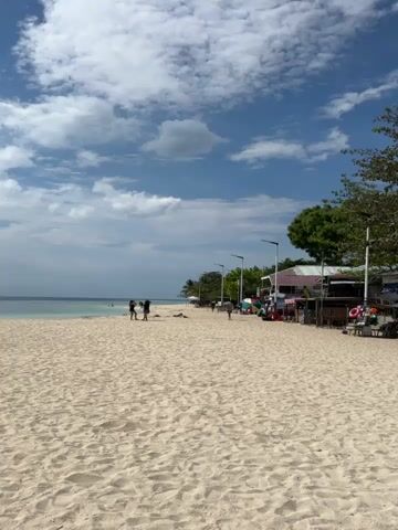 A video captures a serene beach scene with golden sand and clear blue water. Several people are scattered across the beach, some walking, others relaxing. The sky is dotted with fluffy white clouds against a vibrant blue backdrop. Trees line the edge of the beach, providing a touch of green against the sandy expanse. Small stalls with colorful displays are visible along the shore, suggesting a vibrant local atmosphere. The gentle lapping of waves against the shore creates a tranquil ambiance.