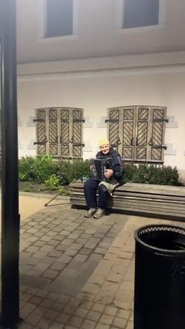 A man sits on a wooden bench playing an accordion. He is wearing a light-colored hat, a dark jacket, dark pants, and sneakers. A cane leans against the bench next to him. He is in front of a building with faux window shutters and a manicured hedge. The ground is paved with brick-like stones. A dark pole is in the foreground on the left side of the frame, and a dark trash can is partially visible on the right. The lighting suggests it is dusk or nighttime.