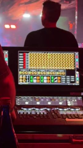 The video captures an energetic night scene at a live music event or club, likely a concert or festival. In the foreground, an individual is operating a complex digital control panel. A laptop screen displays a grid of illuminated yellow and green lights, orange circles, and faders, suggesting a lighting or visual effects control interface. Below the laptop, a physical mixing board with numerous buttons and faders is visible. A blue bottle is also partially visible on the left.

In the background, a person is silhouetted against bright stage lights, appearing to be on a stage and facing away from the camera. The entire venue is bathed in intense red and orange lighting, creating a vibrant and dramatic atmosphere. Red and orange fabric strips are draped decoratively from the ceiling, contributing to the event's aesthetic. The ambient lighting indicates it is nighttime. Upbeat electronic music plays throughout the clip. The location context of Indio, United States of America, suggests a large-scale event. No descriptive text is clearly legible on the screens or equipment beyond what appears to be common interface labels.