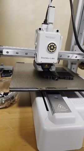 A white Bambu Lab 3D printer, with a print head adorned with a circular array of yellow and black lights, is actively printing a dark object on a textured brown print bed. The print head moves back and forth, extruding material onto the object. The print bed is mounted on a sliding tray, and the printer's base is white with silver accents. A small, clear bowl containing black and white plastic components is visible to the left of the print bed. The background is a plain wall.