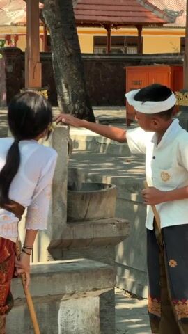 A young boy and girl in traditional Balinese clothing are interacting outdoors. The girl is wearing a white top with decorative trim and a patterned sarong. She is bending over, holding a wooden stick. The boy is wearing a white shirt with a logo, a white headband, and a patterned sarong. He is standing and holding a wooden stick. He is reaching out to touch the girl's hair or head. They are in an area with stone structures and what appears to be a tree. In the background, there is a building with a red roof and a sign that says "TOURIST INFORMATION". The scene is lit by sunlight, casting shadows. The overall impression is one of children engaged in a playful or helpful activity in a culturally significant setting.