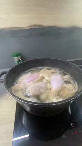This media file depicts a close-up shot of a black, non-stick pot filled with liquid and various ingredients, likely food, simmering on a black induction stovetop. The pot is centered in the frame. Inside the pot, several pieces of what appear to be raw poultry, possibly chicken legs due to their shape and pinkish-white color, are submerged in a cloudy, yellowish broth. Bubbles indicate that the contents are actively cooking. The pot has two black handles.

To the left of the pot, a green-capped spice jar is visible. The label on the jar is partially obscured but the visible text reads "ARTISAN" and "AGRO," suggesting it contains some kind of herb or spice blend.

The background features a textured wall, with the upper portion appearing to be a dark grey, possibly a backsplash, and the lower portion a lighter, wood-grain pattern. The scene is brightly lit, suggesting it is daytime or the area is well-illuminated, but no specific time of day or weather conditions can be inferred. The setting appears to be a kitchen, given the presence of the stovetop and the cooking pot. There are no people visible in the frame. The overall activity is food preparation, specifically the simmering or boiling of poultry. No emotions are conveyed as there are no human subjects. The only discernible text is on the spice jar. The location context of Beş-Küŋgöy, Kyrgyzstan, is provided separately and is not directly inferable from the visual content of the image itself.