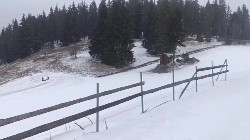 The video shows a snowy landscape with a wooden fence in the foreground. Behind the fence, there is a flat, snow-covered area with a few people. Some appear to be skiing or snowboarding. In the middle ground, there is a building with a red roof and a few vehicles. A road runs along the edge of the snowy area, with buildings on the other side. The background is filled with tall evergreen trees on a hillside. The sky is overcast, and the overall impression is of a winter scene in a mountainous region.