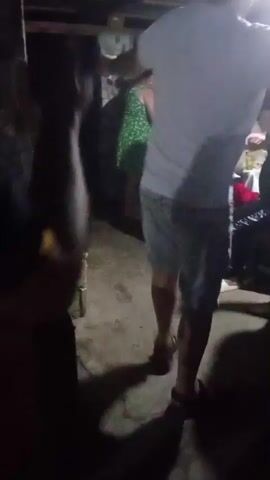 A man in a white t-shirt and grey shorts dances in a dimly lit, open-air space with a concrete floor. He is facing away from the camera. To his right, a woman in a teal shirt and a child in a red and white outfit are seated. Other people are visible in the background, some partially obscured by shadows. The environment appears to be an outdoor gathering or celebration. The man is actively moving his arms and body to the music.