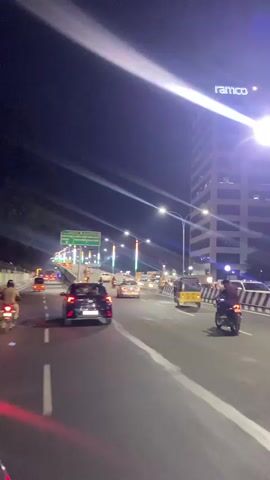 The media file depicts a nighttime scene of a busy city street, likely in Chennai, India. The camera is mounted on a moving vehicle, giving a first-person perspective of the road ahead. A multi-lane road is visible, with several cars, a motorcycle, and an auto-rickshaw navigating through traffic. Streetlights illuminate the road, and colorful lights, possibly festive decorations, adorn some of the lampposts. A tall, modern building with the "ramco" logo illuminated on its top section stands prominently on the right side of the frame. A green directional signpost is also visible, indicating "ADYAR" and "RAJIV GANDHI SALAI (OMR)". The atmosphere appears dynamic and urban, with the glow of artificial lights against the dark night sky. The time of day is clearly night, and there's no indication of specific weather conditions other than the lights reflecting on the wet-looking road surface in some areas. The overall impression is one of a vibrant, active city at night.