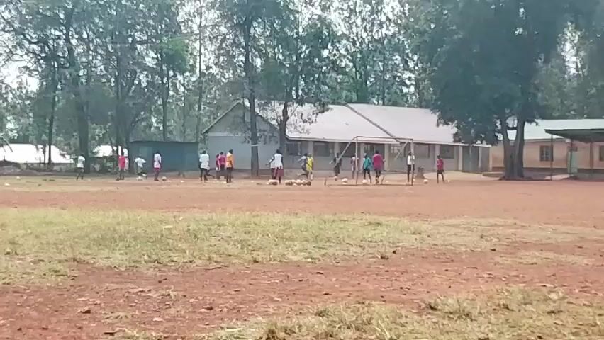 A group of approximately 15 individuals, likely youths or young adults, are engaged in what appears to be a soccer practice on a dirt field. They are scattered around the field, most near the goal post, kicking or standing around soccer balls. The individuals are wearing various colored shirts, including pink, white, and yellow. A building with a long, slanted roof is visible in the background behind the goal post. There are other buildings and trees on the periphery of the field, creating a somewhat rural setting. The foreground shows a mix of brown dirt and patches of dry grass. The weather seems overcast.