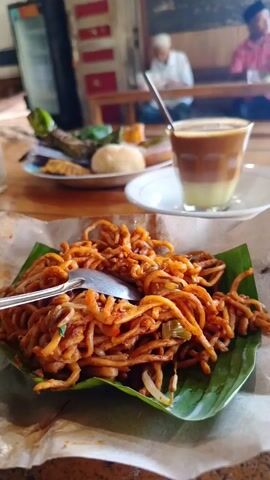 The video shows a plate of mie goreng (fried noodles) served on a banana leaf, with a spoon on top. Behind it, there is a glass of coffee and a plate with various snacks and side dishes. In the background, there are people sitting at a table in what appears to be a restaurant or cafe. A young boy walks past the seated customers. The background also features a refrigerator and a blackboard. The scene is brightly lit, suggesting it's daytime.