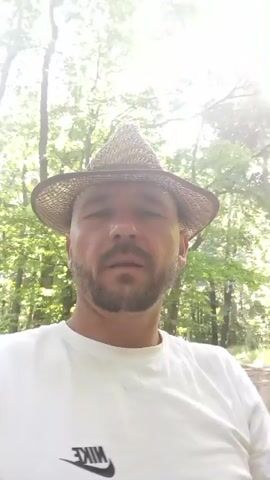 This is a short video clip featuring a man wearing a straw hat and a white t-shirt with a Nike logo. He is outdoors, in a setting that appears to be a forest or wooded area, with sunlight filtering through the trees. The man is looking directly at the camera and smiling, with a beard and short hair. The background is blurred, suggesting movement, possibly from the camera operator walking or running. The scene has a bright, daytime feel, with lush green foliage visible. The man's expression conveys happiness and a relaxed, enjoyable mood. There is no discernible event taking place other than the man possibly recording himself while on a walk or hike. The visible text is the "NIKE" logo on his t-shirt, which is reversed due to the selfie-style recording. The overall impression is one of a cheerful person enjoying nature.