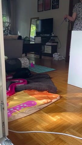 The video shows a living room with a person in a floral dress standing on the right side of the frame, holding a blue rod. The rod is extended towards the left side of the frame, where a black cat is lying on a blanket. The cat appears to be playing with a pink toy. The room is furnished with a sofa, a coffee table, a television, and various decorative items. The floor is made of wooden planks, and sunlight streams in from a window, casting shadows on the floor. There is a white board or easel on the far right of the frame.