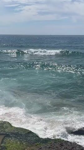 The video shows a body of water with waves. The water is a deep blue color in the distance, transitioning to a turquoise and then white as the waves crash onto a rocky shore in the foreground. The sky above is partly cloudy. There is no evidence of people or any specific objects apart from the natural elements of the sea, rocks, and sky. The waves are the primary action, with white foam and spray visible as they break. The overall environment is a coastal scene under a bright sky.
