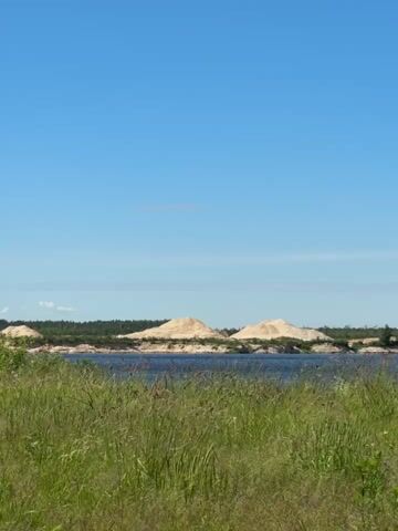 This video shows a landscape under a clear blue sky. In the foreground, there is tall green grass. In the middle ground, there is a body of water with a dark blue surface. Across the water, there is a sandy bank with several large, pale sand dunes. A line of trees forms a darker, lower horizon behind the sand dunes. The scene is calm and natural, with no people or discernible activity.