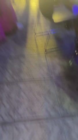 The video shows two costumed individuals in a public space at night. The person in the foreground wears pink pants, white shoes, and a headdress with sunglasses over a blue face. They are holding a staff. Behind them, another person is wearing a black robe with a cross and holding a decorated box above their head. The background includes people, streetlights, and buildings, suggesting an outdoor event or celebration.