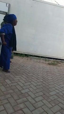 A brief video depicts two women walking on a paved, herringbone brick-patterned surface, with a large white trailer in the background. One woman, dressed in a blue patterned outfit with a matching headwrap, is using a crutch to assist her walking. The other woman, wearing a patterned dress in earth tones, walks beside her. Both women are facing away from the camera and walking towards the right. The setting appears to be outdoors.