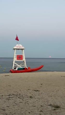 The video shows a beach scene at dusk. A lifeguard tower stands on the sand near the water's edge. A red rescue boat is docked beside the tower. A red flag flies from the top of the tower. The sun is setting over the ocean, casting a pinkish glow on the horizon. The water is calm, and there are some yellow buoys in the distance. The sand appears to be fine and light brown. The sky is a pale blue-gray.