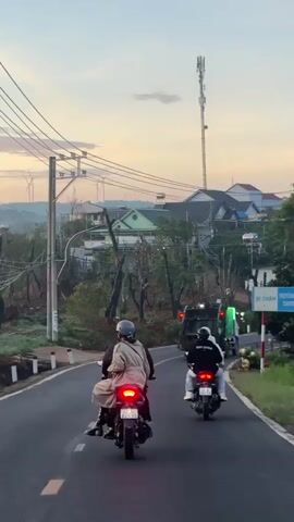Two people on a motorcycle are driving down a paved road. The person in front is wearing a beige coat and a helmet, and the person behind is wearing a black hoodie and jeans. Another motorcycle is driving ahead of them, with two people on it. The person in front is wearing a black hoodie and the person behind is wearing a white hoodie. In the background, there are houses, trees, and a cell phone tower. The sky is a light orange, suggesting it is either sunrise or sunset.