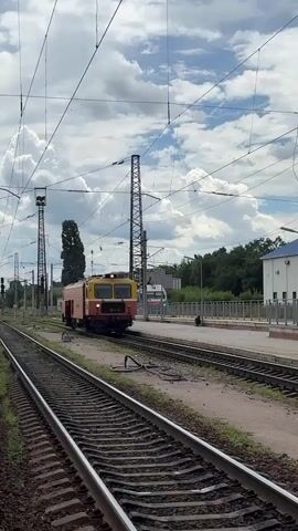 A train with orange and yellow cars moves on railroad tracks. The train is in motion and appears to be approaching or passing by the viewer. The sky is cloudy with blue patches visible. Electrical wires run overhead, supported by tall poles. A building, possibly a station, is visible in the background on the right side, along with trees and foliage. There are multiple sets of parallel railroad tracks. The scene appears to be an outdoor railway environment.