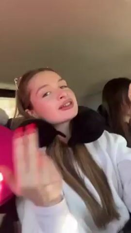 The video shows a young woman with light skin, long blonde hair pulled back in a ponytail, and bright red nails. She is wearing a white hooded sweatshirt and a black travel pillow around her neck. She is in the passenger seat of a car, talking and gesturing with her hands. Sunglasses are perched on her head. In the background, another young woman with brown hair, wearing a black jacket, is partially visible. The car interior is visible, and bright sunlight streams in. The woman in the passenger seat seems to be addressing the camera directly.