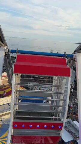 The video is an aerial view from a Ferris wheel looking down on a carnival. The Ferris wheel car is in the foreground, with a red and blue top. Below the Ferris wheel car, there is a yellow and red striped tent and various carnival rides and booths. There are many people walking around the carnival grounds. In the background, there is a body of water and buildings. The sky is blue with some clouds.