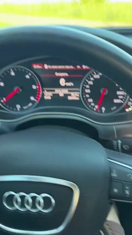 The video shows a first-person view from inside a car. The driver's hands are on the steering wheel, which has the Audi logo. The car is driving on a dirt road through a rural area with green fields and trees on either side. The sky is blue with some clouds, suggesting it is daytime, likely late afternoon or early evening due to the lighting.

The dashboard instruments are visible, including a speedometer displaying a speed of 8 km/h at the beginning of the clip, which increases to 9 km/h. The display also shows the date as 24.5.2026 and other car information. The car appears to be an Audi, given the logo on the steering wheel.

The setting is a rural landscape in Dovhe, Ukraine, characterized by unpaved roads and open fields. The mood is calm and relaxed, as the car is moving at a slow pace through a peaceful natural environment. There are no people visible besides the driver's hands. The overall impression is one of a leisurely drive through the Ukrainian countryside.