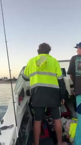 The video shows a man in a bright yellow high-visibility jacket and black shorts standing at the helm of a motorboat moving on the water. A fishing rod is visible, secured near the boat's console. The camera then shifts to show the open water ahead, under a clear sky with a horizon tinged by the colors of sunrise or sunset. The water is moderately choppy, with small waves visible. In the distance, a low landmass with trees can be seen.