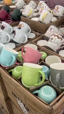 The video shows a close-up view of a large display of ceramic mugs in cardboard boxes. The mugs are arranged in rows and stacked within the boxes. They come in various colors, including pastel shades like light blue, green, pink, and yellow, as well as darker colors like maroon and dark green. Many of the mugs have a ribbed or textured surface. Some mugs have simple designs or illustrations, such as cats or coffee-related motifs. In some of the boxes, the mugs are accompanied by matching saucers. The lighting suggests an indoor setting, possibly a store or warehouse, with overhead lighting casting shadows on the items. The camera angle is slightly tilted, and the focus shifts between different sections of the display, highlighting the variety of mugs. There are no people visible in the frame. The overall impression is of a retail inventory or a display setup for selling mugs.