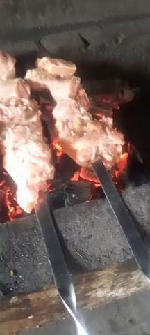 The video shows chicken pieces on skewers being grilled over hot coals. The chicken appears to be raw or lightly cooked, with some seasoning visible. The coals are glowing red, indicating intense heat. The camera is positioned close to the grill, providing a detailed view of the cooking process. The background is dark and indistinct, suggesting an outdoor or dimly lit setting.