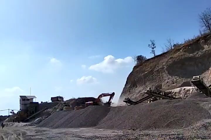 A quarry is depicted under a clear blue sky with scattered clouds. A steep, rocky hill dominates the right side of the frame, with several conveyor belts leading down from its upper reaches. Piles of gravel and crushed stone are visible at various stages of processing. In the foreground, a large mound of dark gray gravel sits prominently. To the left, a small building with a white structure and a red roof is situated, adjacent to a white pillar or mast. Several heavy machinery vehicles, including excavators and trucks, are present at the quarry. One red excavator is actively digging or loading material. A person is walking in the lower left corner of the frame. The overall scene suggests an active industrial site for material extraction and processing.