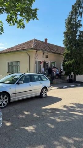 The video shows a one-story building with a red tile roof and light-colored walls, seemingly an office or public building, as there are people gathered on its front steps. The building has a chimney and a small entrance area adorned with flags or banners. Several people are standing around the entrance, and some are on the steps. A silver BMW station wagon is parked in front of the building. In the lower left corner of the frame, there is a large bottle of water on the ground. The building is shaded by trees and the sky is blue with some clouds.