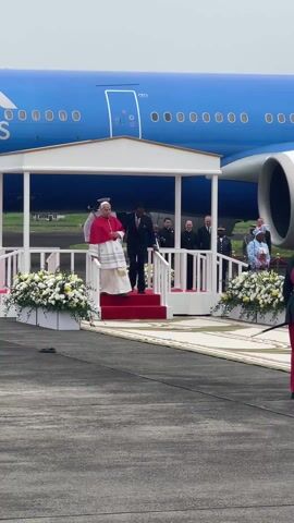 The video captures a formal arrival or departure ceremony on an airport tarmac, likely in Malabo, Equatorial Guinea. The central figure, an older man dressed in a white cassock with a red cape, is seen descending red-carpeted stairs from a white ceremonial platform. He is escorted by a man in a dark suit.

Several other men in suits and uniforms, including what appears to be a military officer in a white uniform, are visible on the platform. On the right side of the frame, a person in a red and dark green military uniform with a sword stands as part of a potential honor guard.

In the background, a large blue passenger airplane, possibly a wide-body jet, dominates the scene. The aircraft features rows of circular windows, a prominent engine on its right wing, and partial white text including the letter "S" and numbers "0000000" on its fuselage.

The ceremonial platform is decorated with elegant arrangements of white and yellow flowers on either side of the stairs. The ground is a grey concrete tarmac. The sky appears overcast, suggesting a cloudy daytime event. The audio indicates cheers and a ceremonial fanfare, highlighting the formal and significant nature of the occasion.