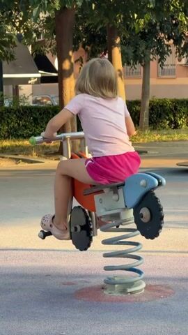 A young girl with blonde hair, wearing a pink t-shirt and pink shorts, is riding a spring-loaded toy motorcycle. She is on a playground, and the sun is shining, casting long shadows. The girl is holding onto the handlebars of the motorcycle, and her legs are positioned as if she is riding. The toy motorcycle is blue and orange with black wheels and a grey spring. It's a sunny day, and the girl appears to be enjoying herself. In the background, there are trees, buildings, and other playground equipment. The ground of the playground is paved with a speckled surface. There is another child in the background wearing a bright yellow shirt. The scene takes place in Benimàmet-beniferri, Spain. There is no visible text in the media file.