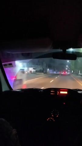 The video shows the view from inside a car at night, looking out the windshield. The road ahead is illuminated by streetlights and the car's headlights. Traffic lights are visible, as are other vehicles on the road. The dashboard of the car is visible in the foreground, with illuminated gauges and a digital display. The overall scene depicts driving at night on a paved road lined with trees and buildings.