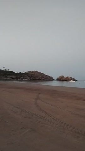 This media file shows a view of a beach in Águilas, Spain. The foreground is dominated by a sandy beach with tire tracks visible. In the background, there are two rocky islets in the sea. Several boats are present in the water, including a white speedboat near the islets and another white boat further out to sea. The sky is overcast, suggesting it is either early morning or late evening. There are no people visible in the media file. The overall mood is peaceful and serene.