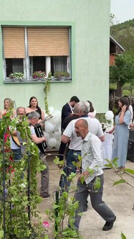 The video captures a group of people gathered in what appears to be a garden or backyard setting, enjoying live music and interacting with one another. Musicians are playing instruments, including a guitar, accordion, and violin. Several other individuals are standing around, seemingly part of the group, with some interacting with each other through conversation and gestures like hugging. The location features a light green house with closed blinds and flower boxes in the window. Decorations like white balloons and a small white table add a festive atmosphere. In the foreground, a trellis covered with green vines adds a touch of nature to the scene. Overall, it seems to be a lively, social gathering taking place outdoors.
