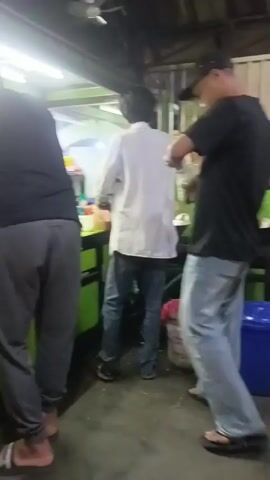 The video shows the interior of a food stall. Three individuals are present. One individual, wearing a black t-shirt and light blue flared pants, stands facing towards the right, holding a plastic bag of food. Another individual, wearing a white and blue striped long-sleeved shirt and blue jeans, is facing away from the camera and appears to be working at a stove. A third individual, wearing a black t-shirt and grey sweatpants, is partially visible on the left side of the frame, facing towards the stall. The stall is equipped with a stove, cooking utensils, and bright green countertops. A blue bucket and a red-lidded trash can are visible on the floor. The lighting suggests it is nighttime or an indoor setting.