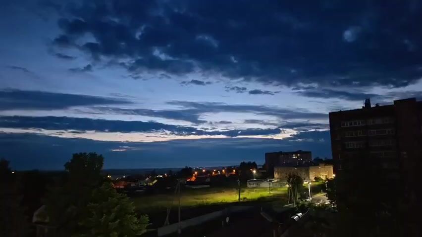 The video shows a nighttime aerial view of a town or city at dusk. The sky is dark blue with streaks of clouds. In the foreground, there are trees and buildings. In the midground, there is a field with some scattered lights. In the background, there are more buildings and lights of the town. The overall mood is calm and serene.