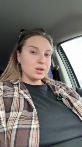 A young woman with long blonde hair and hoop earrings is sitting in the driver's seat of a car. She is wearing a black t-shirt and a brown, white, and red plaid flannel shirt. Sunglasses are perched on her head. She is looking at the camera and lip-syncing to a song that is playing. She then turns her head slightly and looks through the driver's side window, smiling. The background shows the car's interior and a blurred view of the outside through the window.