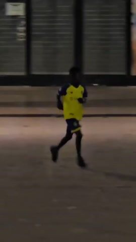 A young boy in a yellow and blue soccer uniform with the number 7 on the back is playing soccer. He is running with the ball, kicking it, and then another boy in a white tank top joins him to play. The scene appears to be outdoors at dusk, possibly in an urban area, with storefronts in the background.
