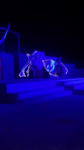 The video shows two performers on a stage at night, illuminated by blue lights. They are wearing costumes with large, light-up wings that create a beautiful spectacle as they move them in circular motions. The stage is simple, featuring steps and a backdrop, with a skeletal frame structure visible on the left. The overall scene has a dark and mysterious atmosphere, with the focus entirely on the dancers and their illuminated wings.