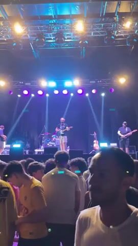 The video shows a concert with a band performing on stage. The stage is lit with blue and purple lights. There are four band members on stage: one playing guitar and singing, one playing guitar, one playing drums, and one playing bass guitar. The audience is standing in front of the stage, watching the performance. The video is shaky and out of focus, suggesting it was taken by someone in the crowd. The sound of the music is muffled.
