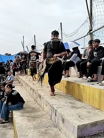 A person wearing black clothing, including a shirt, pants, and a face covering that extends up to their eyes, is walking down concrete steps while carrying a large, spiky, and what appears to be a dried or preserved animal in their left arm. In their right hand, they are holding a black bucket. The person is barefoot. Several spectators are seated on bleachers behind them, some looking towards the person and the item they are carrying. A net is visible in the background, possibly part of a sports field. There is a banner behind the spectators with text in a non-English language. The environment appears to be outdoors under an overcast sky.
