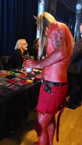A man dressed as a devil stands at a table, holding a white mug. He has long blonde hair, a red complexion, horns, and a tail. He's wearing red shorts and a black belt. The table is covered with merchandise, including CDs and small decorative items. Behind the table, two women sit in chairs and converse. Another man in a suit stands to the side, talking to someone out of frame. The setting appears to be an event or convention, with dark curtains in the background and other attendees visible.