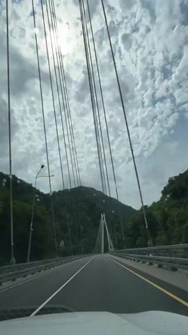 This video captures a view from inside a vehicle driving across a modern cable-stayed bridge. The bridge spans a deep, lush green valley, with steep, tree-covered mountains on either side. The sun is shining brightly through a sky filled with dramatic, puffy white clouds, suggesting it's daytime. The road is paved and marked with white and yellow lines, indicating a highway or major road. The bridge itself features a distinctive white pylon that supports the numerous cables extending downwards to hold up the deck. On the sides of the bridge, there are metal guardrails and streetlights. A road sign is visible on the left side of the road. The overall impression is one of a scenic drive through a mountainous landscape, with the impressive engineering of the bridge forming a striking contrast with the natural environment. The video appears to be filmed from the front passenger seat of a car, as the dashboard is partially visible at the bottom of the frame. There is no discernible text within the video frames.