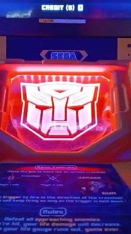 The camera shows a close-up view of a "Transformers" arcade game. The game's screen, lit up with the Autobot logo, switches between red and blue. A "SEGA" logo is visible at the top center of the machine. Below the screen is a panel explaining the game's basic controls and rules. The panel mentions using the gun to move the on-screen crosshair and firing with the trigger. The rules state that players must defeat all approaching enemies, and losing life will result in game over. The video is stationary.