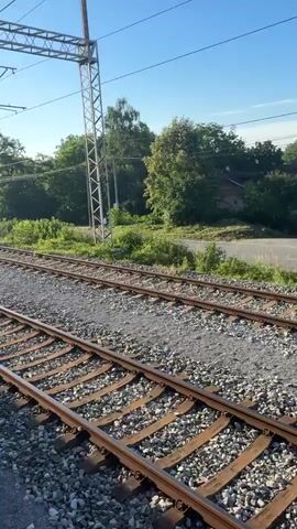 The video shows a view from a train moving along a double set of train tracks. The tracks are made of gravel and steel rails. Overhead, power lines stretch across the sky, supported by metal poles. Trees and greenery line the sides of the tracks, with a clear blue sky above. In the distance, a few white vehicles are visible on a road. The train appears to be moving at a steady pace, with the landscape passing by smoothly. There are no people visible in the video. The scene is tranquil and depicts a typical train journey through a rural or suburban area during daylight hours.