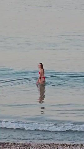 A young woman wearing a red bikini is standing in shallow ocean water. The water is calm with small waves breaking near the shore in the foreground. The woman is facing away from the camera, looking to her left. She has long brown hair and appears to be posing. The background consists of the ocean stretching to the horizon, with a pale, overcast sky. The shoreline in the foreground is composed of pebbles.