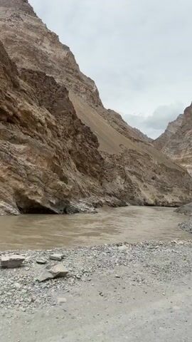A vehicle drives along a gravel road that runs beside a wide, murky river. The river flows through a narrow canyon with steep, rocky sides. The sky is cloudy. The terrain is arid and mountainous.