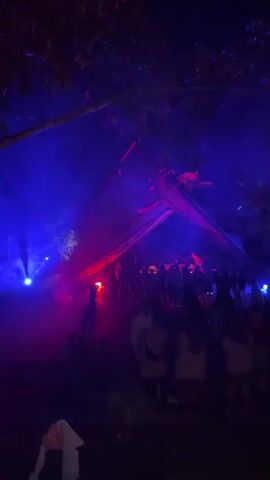 The video captures an outdoor nighttime event, likely a party or festival, set in Penebel, Indonesia. Numerous people are gathered, with some standing and others seated, under a canopy of trees. The scene is dramatically lit with strong, pulsating blue and red lights, which highlight a smoky or foggy atmosphere. A large, dark, triangular structure, possibly a stage or a tent, is visible in the background, also illuminated by the colorful lights. Upbeat electronic music plays throughout the clip, contributing to the lively ambiance. No discernible text is visible.